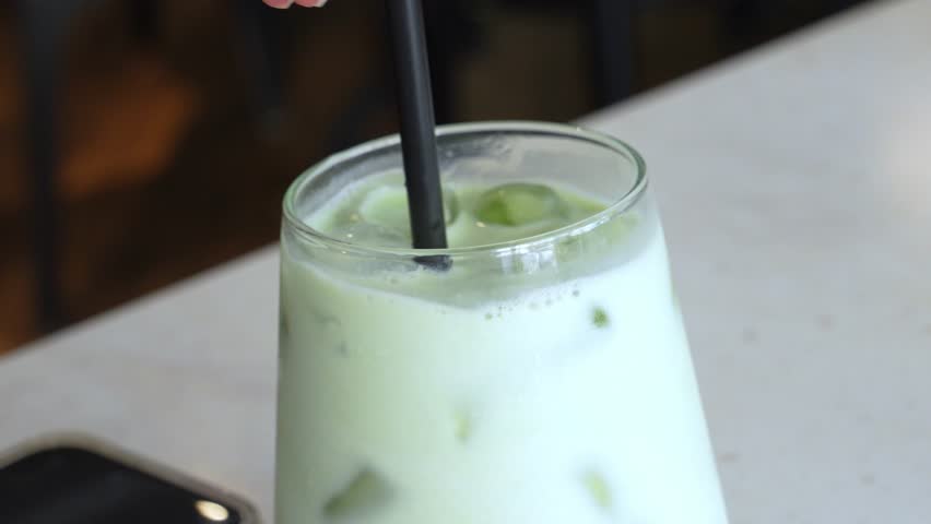 A close-up of a green matcha cold latte in a cup. The latte features a heart-shaped foam design on top. A dessert is partially visible in the background.