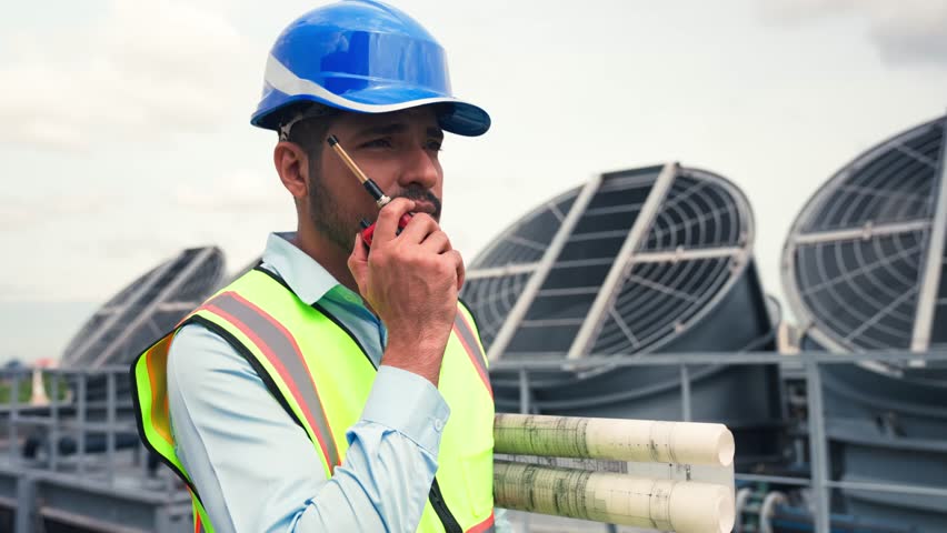 Engineering people with Asian male engineer using walkie talkie on rooftop to assign jobs while inspecting HVAC cooling tower system.