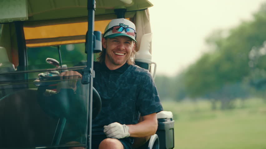 Happy male golfer sits in the driver seat of a golf cart, smiling and laughing in slow motion with one hand on the steering wheel. The clip captures a candid and joyful golf course moment.