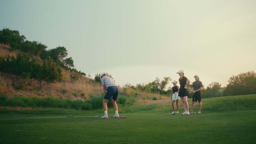 Golf foursome gathers on a par three as a male golfer hits his tee shot at sunset. The group watches intently in golden hour light, capturing a calm and scenic golf moment with shared focus