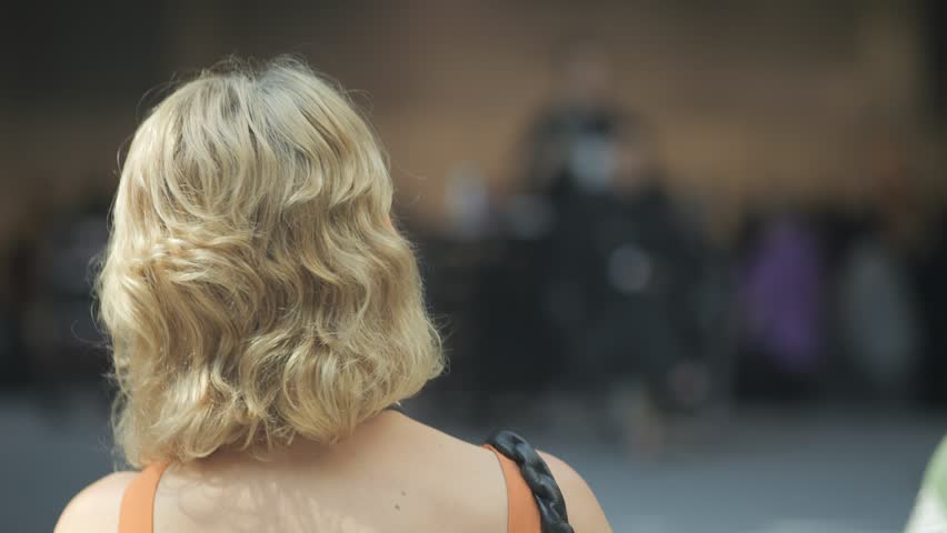 Woman with blonde hair is standing in front of a building. She is wearing an orange tank top and a black purse