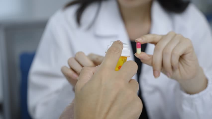 Medical professional in white coat discussing pharmaceutical treatment, holding medication bottle and pill while providing healthcare advice to patient