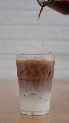 4K footage vertical video Front view close-up iced milk in a clear plastic cup being topped with a fresh pour of coffee, swirling blend of dark coffee and creamy milk.