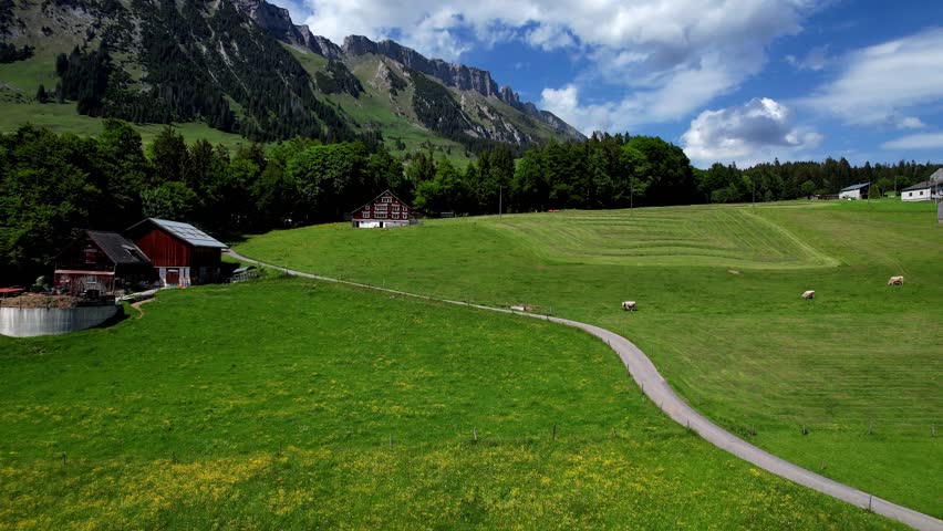 A 4K drone shot of a traditional Swiss farmhouse at the edge of a lush green valley. Surrounded by forest, alpine meadows, and dramatic mountain ridges under a partly cloudy sky in Switzerland.