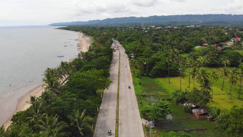 Drone view of winding coastal road beside sandy beach lined with palm trees and lush greenery in Philippines. Gentle waves reach shore as scattered boats float near tropical shoreline.