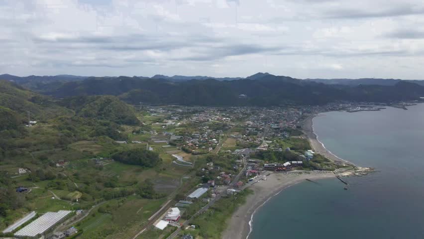 Aerial drone shot of Kamogawa City showing scenic shoreline, lush hills, beachside houses, farmland, and peaceful oceanfront landscape under soft clouds in calm daylight