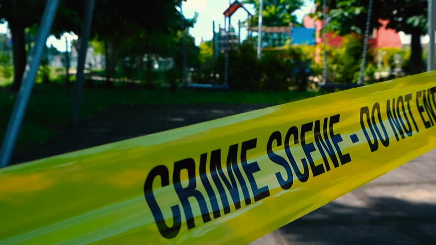 Static view of yellow crime scene tape, do not enter in foreground, sunny day. In blurred background closed playground park, empty moving swings. Crime against child, kid or children or missing person