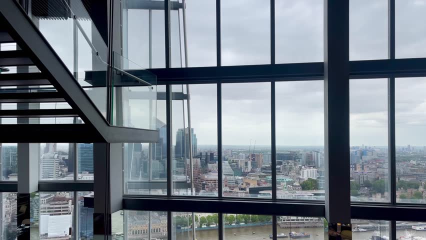London cityscape view through large glass windows from a high-rise interior overlooking buildings and rooftops on a cloudy day, slow motion