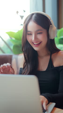 Happy asian woman using laptop with wireless headphones at modern coffee shop, reusable cup on table – digital lifestyle, eco-conscious living and urban comfort