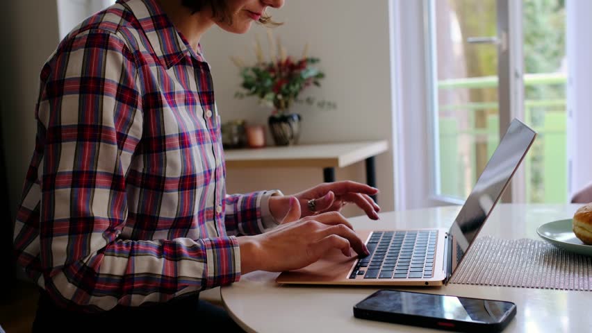Smiling adult woman in works from home in sunny kitchen, typing on laptop, enjoying work-life balance, singing joyfully, feeling relaxed and inspired by remote job freedom.