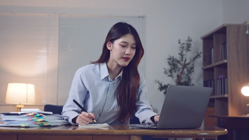 Professional businesswoman working at desk, taking notes with laptop, displaying focused productivity and positive demeanor in corporate environment