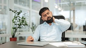 Professional Indian businessman seated at an office desk, dressed in a light shirt, experiencing acute neck pain and discomfort. Shows fatigue from a sedentary job, stretching for relief. - Powered by Shutterstock - Get 15% off with code: PIKWIZARD15