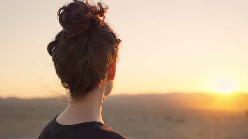 Young woman with back to camera looking at warm orange desert sunset or sunrise. Female traveler or hiker camping outdoors in nature peacefully and calmly standing on windy mountain looking at horizon