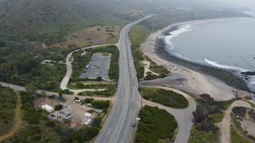 Overhead shot of car driving towards the camera up the Pacific Coast Highway on the California coast. Contrasted by lush greenery and waves crashing onto the beach below. 4K drone road trip shot.