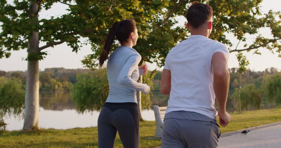 Sporty young couple jogging together along park path during sunrise. Back view of athletic woman and man in sportswear running together, enjoying healthy outdoor activity in nature. Slow motion.