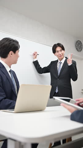 A handsome Japanese businessman wearing a suit giving a presentation at an internal meeting. He stands in front of a whiteboard and speaks. Vertical video.
