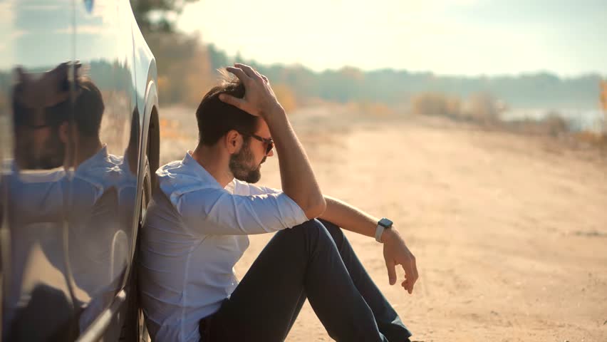 Man sitting alone by roadside with his hand in his hair, leaning against a car. A thoughtful and emotional moment, reflecting solitude, stress, personal contemplation. Burnout tension mental fatigue
