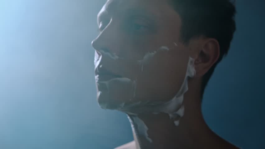 A young man shaves his face with a razor in a steamy bathroom after taking a shower. Close-up cinematic shot with moody blue light