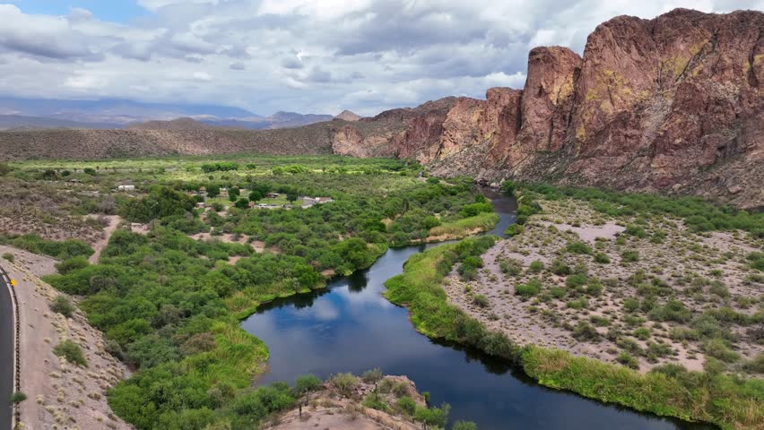 Aerial view of a deep blue river winding through a vibrant green landscape, contrasting with the rugged red rock mountains in the distance, Mesa, Arizona, United States.
