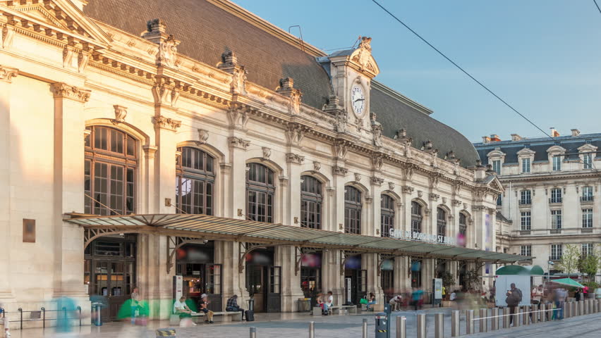 Bordeaux Saint Jean Train Station timelapse, the main railway hub in Aquitaine, France, features a grand entrance with a clock. Trams pass by during sunset, moving shadows on the historic facade