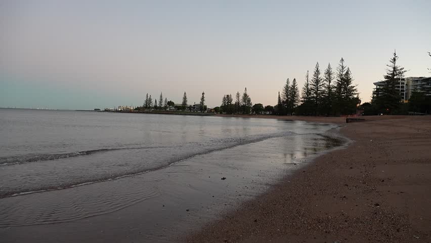 Twilight settles over a calm coastal shoreline in Queensland, Australia. The gentle waves lap against the beach, framed by silhouetted pine trees and soft hues of the fading sky.