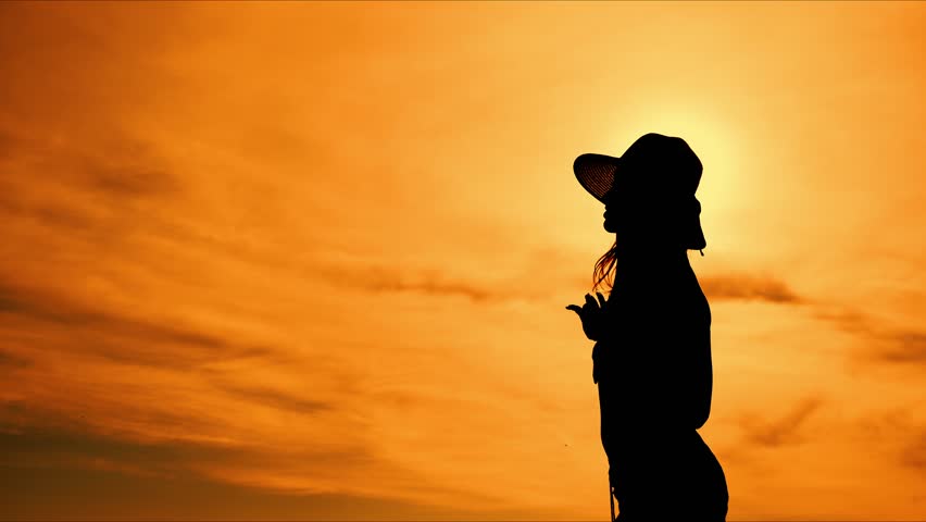 Silhouette of dancing girl in hat at sunset on beach and spreading arms to sides and raising up. Girl tourist on vacation on beach.
