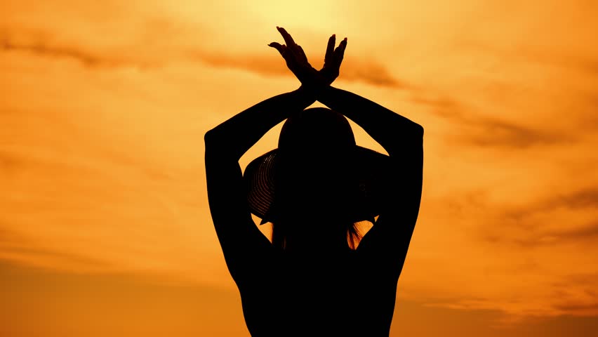 Close-up silhouette of dancing girl in hat at sunset on beach and spreading arms to sides and raising up. Girl tourist on vacation on beach.