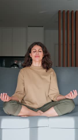 Relaxed young indian woman sitting on couch, meditating with closed eyes in lotus pose. Peaceful latin hispanic or middle eastern brunette girl doing yoga, reduce stress on sofa at home. Vertical