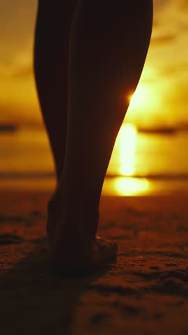 Beautiful sunset or sunrise on beach, closeup view of female legs walking over sand, silhouette against sun. Vertical video with one female figure on coastline, feel happiness, calmness and solitude