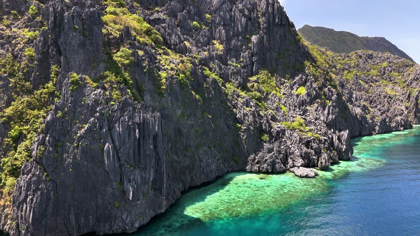 Steep limestone cliffs rise above clear turquoise waters along the rugged coastline of Tapiutan Island, north of Talisay Beach in the Philippines, surrounded by lush greenery under a bright sky