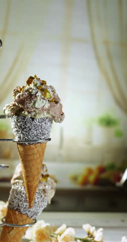 A close-up of a hand gently removing a pistachio-topped vanilla chocolate ice cream cone from a display holder, highlighting the creamy texture and coconut-coated cone in a cozy setting.