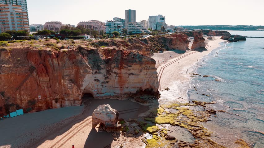 Ocean waves gently caress sandy shores as people stroll along the beach. The rugged cliffs adorned with greenery create a stunning backdrop in Portugal.