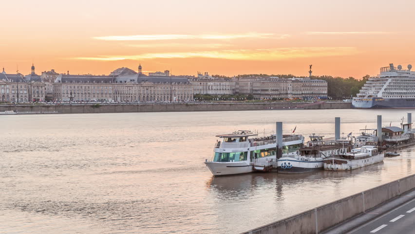 Ship and boats docked along the Garonne River in Bordeaux, France, near the ferry port. Day to night transition timelapse with waterfront reflections, traffic on the street and historic architecture