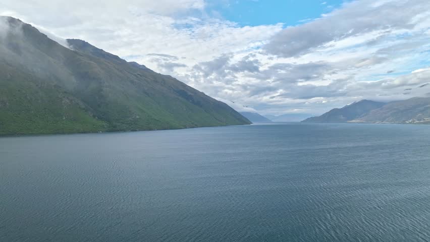 Aerial view of the serene lake embraced by majestic mountains under a cloudy sky, exhibiting a contrast of cool blues and lush greens, Queenstown, Otago Region, New Zealand.