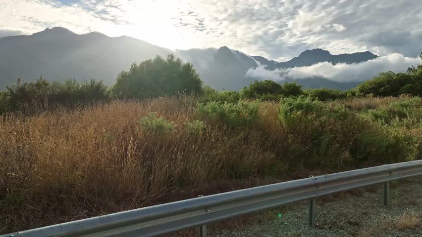 Aerial view of the still water reflecting the sunlight, contrasting with the dark, imposing mountains partially shrouded in clouds, Queenstown, Otago Region, New Zealand.