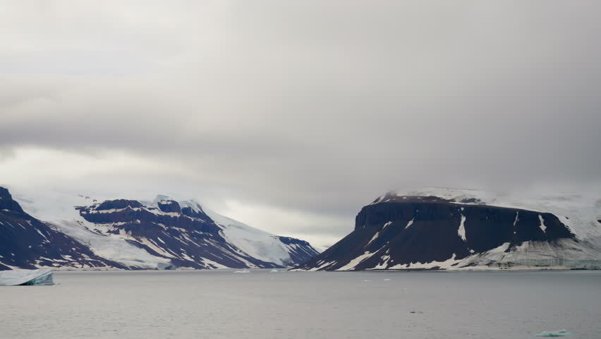 Majestic snow-capped mountains and rugged cliffs rise above an icy Arctic bay, featuring dramatic overcast skies and floating icebergs in a tranquil winter seascape.