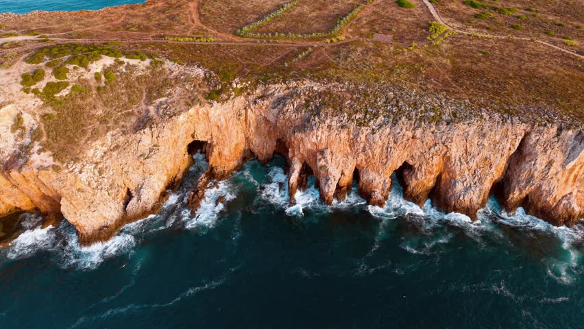 This aerial perspective captures the rugged beauty of Portugal's coastline, highlighting dramatic cliffs and crashing waves under a colorful sunset sky.