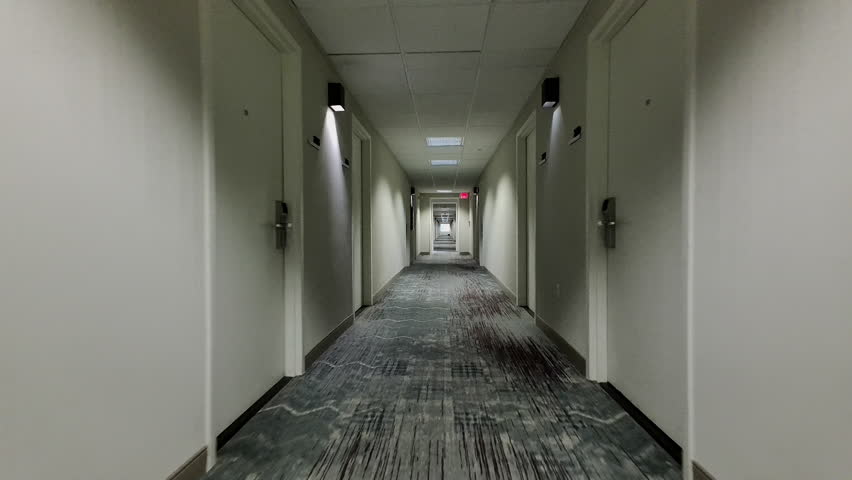 Backward trucking shot down hotel corridor past doors and bland decor.