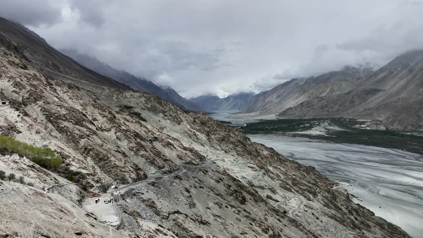 Aerial view of winding road cutting through the rugged, rocky mountains alongside a wide riverbed under a cloudy sky, Ladakh, Ladakh Division, India.