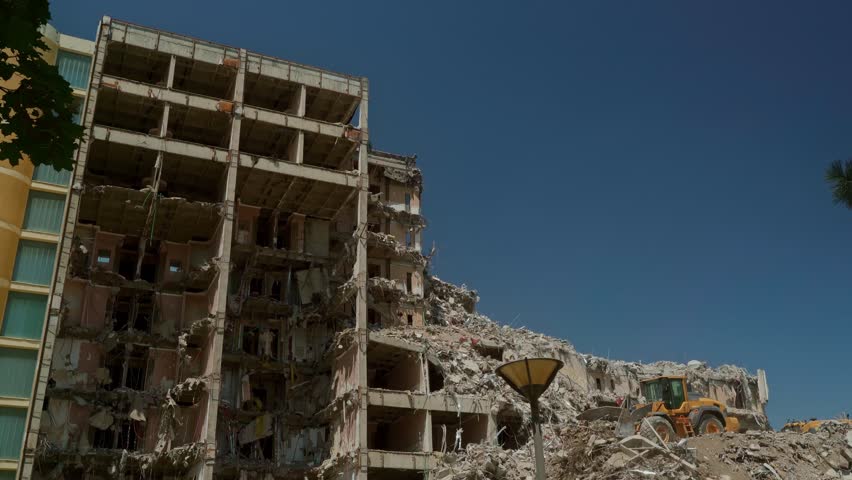Demolition site with a collapsed building, exposed rebar, and a bulldozer working amid concrete rubble and dust. Apartment building destroyed by an earthquake or accident.