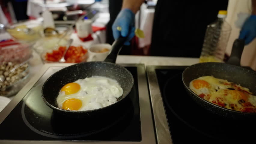 Close-up view of a chef preparing sunny side up eggs and a vegetable omelette in frying pans on a stovetop, focusing on the cooking process in a bustling kitchen setting.