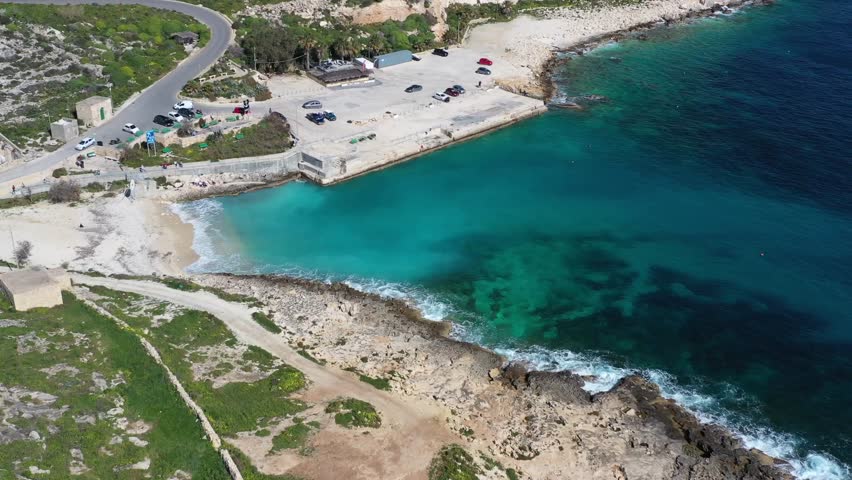 Aerial view of Mgarr ix-Xini Beach, where turquoise waters meet the shore, contrasting with the rugged coastline and the winding road above, Gozo island, Malta.