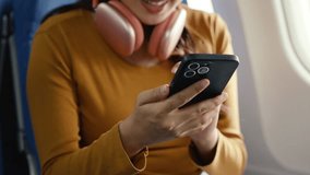 Young woman wearing casual attire and headphones around neck, sitting near airplane window while using smartphone during flight, browsing content with relaxed demeanor - Powered by Shutterstock - Get 15% off with code: PIKWIZARD15