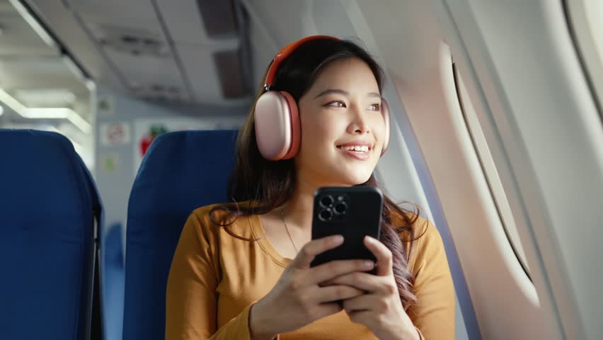 Young female passenger wearing wireless headphones scrolling smartphone while sitting by airplane window, enjoying in flight entertainment during leisure travel