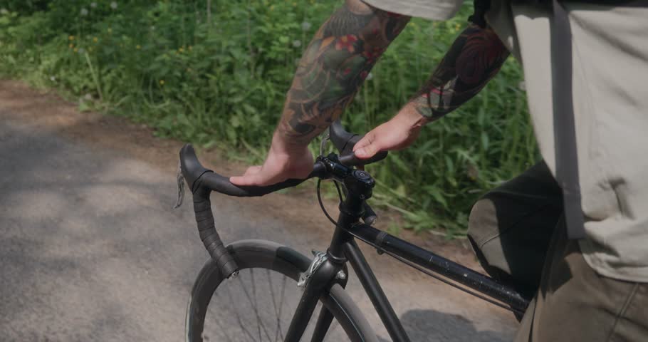Detailed shot of tattooed hands holding the handlebars of a black bike in motion. Forest road, flickering sunlight, and steady grip reflect mindful and active lifestyle. High quality 4k footage
