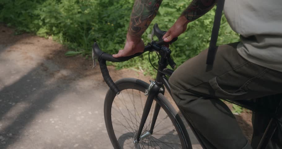 Detailed shot of tattooed hands holding the handlebars of a black bike in motion. Forest road, flickering sunlight, and steady grip reflect mindful and active lifestyle. High quality 4k footage