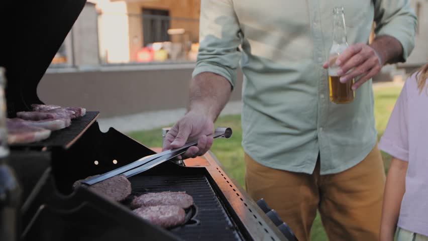 Father grilling hamburgers and drinking beer while his daughter watches him at a backyard barbecue