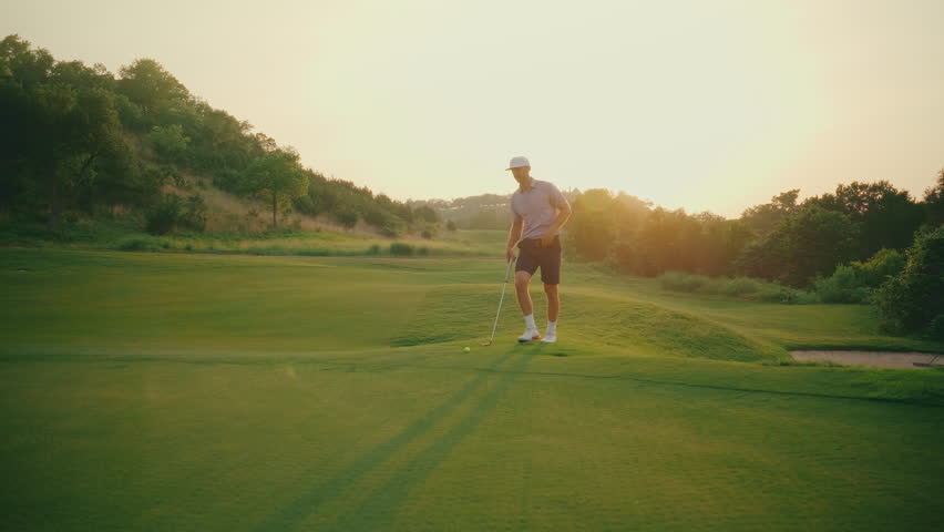 Slow motion walk-up shot of a male golfer putting from the fringe with golden hour sun flares in the frame. A warm, cinematic moment capturing the quiet focus and beauty of evening golf.