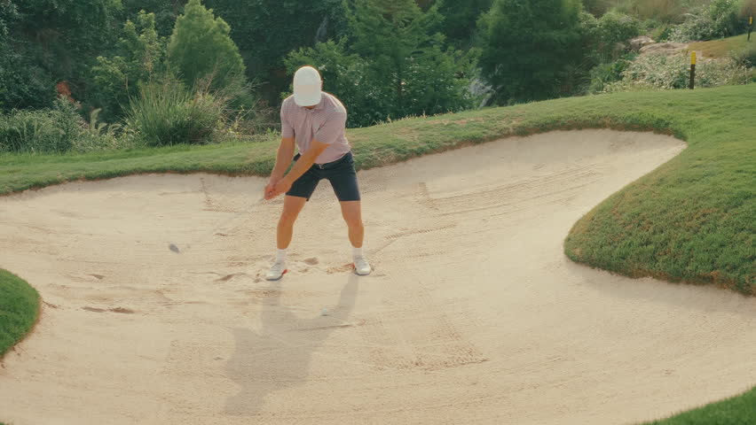 Male golfer hits a textbook bunker shot from a greenside sand trap in slow motion. Clean, smooth swing with sand flying as the ball lifts toward the green. Shot captures perfect short game execution.