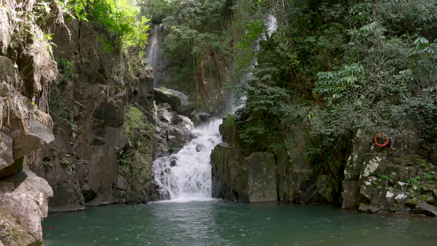 Powerful waterfall cascading between rugged cliffs into emerald pond surrounded by dense forest in tropical national park landscape during peaceful sunny day in Chanthaburi Thailand